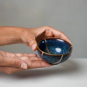 May include: A small, round ceramic bowl featuring a deep blue and black mottled glaze. The bowl displays visible cracks repaired with a gold-coloured lacquer, highlighting the Kintsugi technique with distinct metallic lines across the surface.
