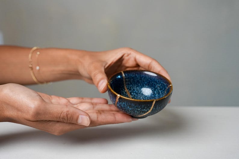 May include: A small, round ceramic bowl featuring a deep blue and black mottled glaze. The bowl displays visible cracks repaired with a gold-coloured lacquer, highlighting the Kintsugi technique with distinct metallic lines across the surface.