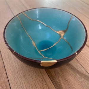 May include: A round ceramic bowl with a vibrant turquoise interior and a dark brown exterior. The bowl features visible cracks repaired with gold-colored lacquer, creating a distinct branching pattern across the inner surface and rim.