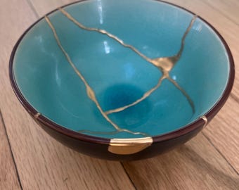 May include: A round ceramic bowl with a vibrant turquoise interior and a dark brown exterior. The bowl features visible cracks repaired with gold-coloured lacquer, creating a distinct branching pattern across the inner surface and rim.