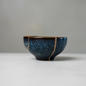 May include: A small ceramic bowl featuring a deep blue and black mottled glaze with a glossy finish. The bowl displays distinct vertical lines of gold-coloured repair material running down its exterior surface, creating a segmented appearance.