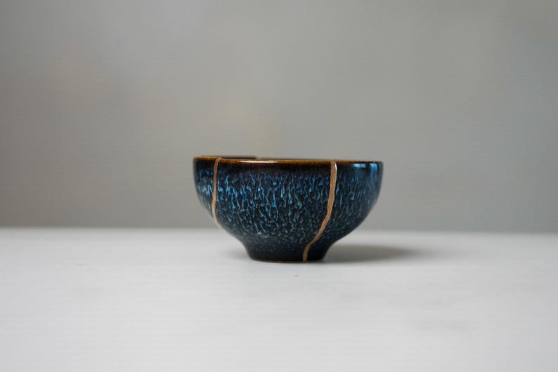 May include: A small ceramic bowl featuring a deep blue and black mottled glaze with a glossy finish. The bowl displays distinct vertical lines of gold-coloured repair material running down its exterior surface, creating a segmented appearance.