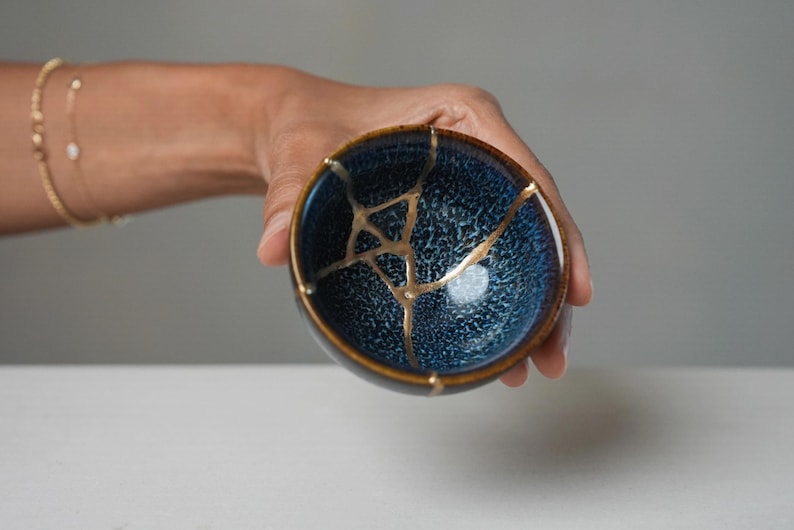 May include: A small, round ceramic bowl featuring a deep blue, textured glaze with a dark brown rim. The bowl displays a prominent kintsugi repair, where the cracks are filled with a distinct, reflective gold-coloured lacquer finish.