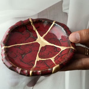 May include: A small, round ceramic dish featuring a deep red glaze with a dark, crackled texture. The surface is repaired with gold-colored lacquer in a kintsugi style, creating distinct lines that connect across the center of the dish.