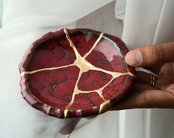 May include: A small, round ceramic dish featuring a deep red glaze with a dark, crackled texture. The surface is repaired with gold-coloured lacquer in a kintsugi style, creating distinct lines that connect across the centre of the dish.