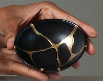May include: A small, oval-shaped bowl with a matte black finish. The surface of the bowl features several prominent, branching cracks that are filled with a smooth, reflective gold-coloured material, creating a distinct contrast against the dark base.