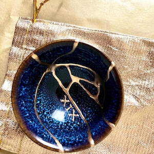 May include: A small, round, deep blue ceramic bowl featuring a textured, mottled surface with a dark brown rim. The bowl is repaired with gold-coloured lacquer in a kintsugi style, forming branching lines and the Roman numeral XIX on the interior.