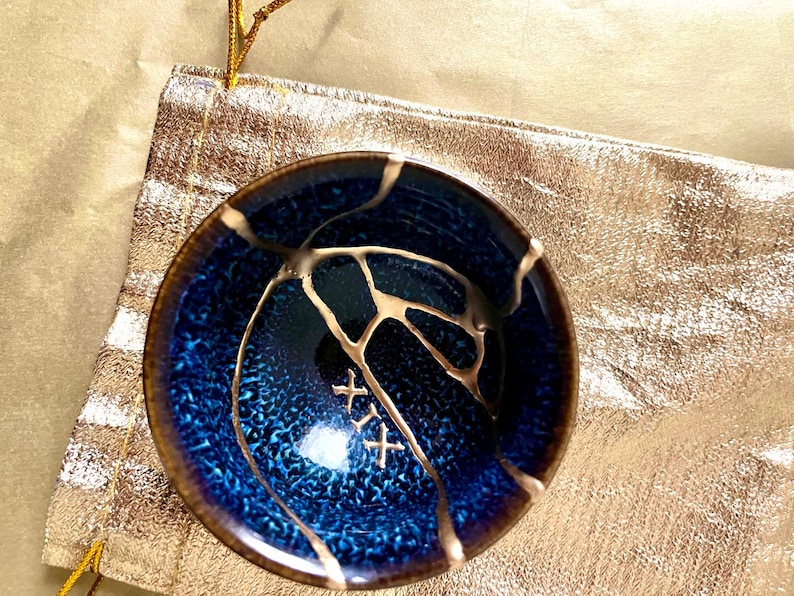 May include: A small, round, deep blue ceramic bowl featuring a textured, mottled surface with a dark brown rim. The bowl is repaired with gold-coloured lacquer in a kintsugi style, forming branching lines and the Roman numeral XIX on the interior.