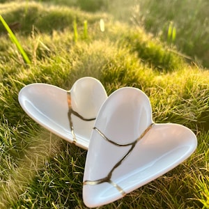 May include: Two white heart-shaped ceramic dishes featuring gold-colored cracks across the surface. The dishes have a smooth, glossy finish and a concave shape, with the gold lines creating a distinct pattern across the white ceramic material.