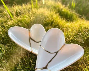 May include: Two white heart-shaped ceramic dishes featuring gold-coloured cracks across the surface. The dishes have a smooth, glossy finish and a concave shape, with the gold lines creating a distinct pattern across the white ceramic material.