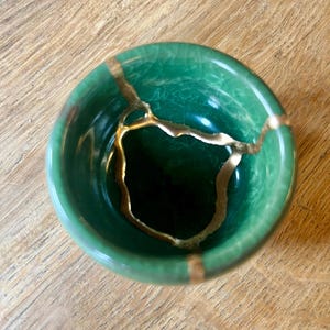 May include: A small, round, emerald green ceramic bowl featuring a visible crackled glaze finish. The bowl is repaired with gold-colored lacquer, highlighting the fractures across the interior surface in a distinct, branching pattern.