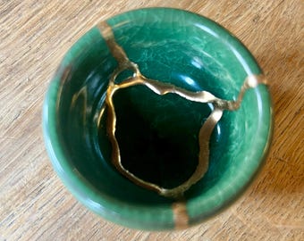 May include: A small, round, emerald green ceramic bowl featuring a visible crackled glaze finish. The bowl is repaired with gold-coloured lacquer, highlighting the fractures across the interior surface in a distinct, branching pattern.