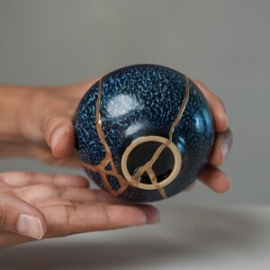 May include: A small, spherical ceramic bowl featuring a deep blue, textured glaze. The surface is marked by distinct, branching cracks repaired with a metallic gold-coloured lacquer, highlighting the breaks across the exterior and the base.