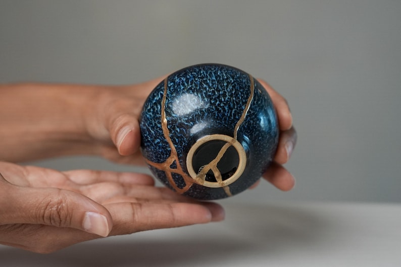 May include: A small, spherical ceramic bowl featuring a deep blue, textured glaze. The surface is marked by distinct, branching cracks repaired with a metallic gold-coloured lacquer, highlighting the breaks across the exterior and the base.