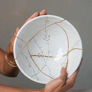 May include: A white ceramic bowl featuring a kintsugi-style repair with gold-colored lines connecting various sections. The interior surface is decorated with delicate, hand-painted light blue and grey floral patterns scattered across the bowl.