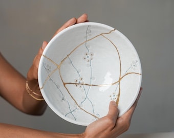 May include: A white ceramic bowl featuring a kintsugi-style repair with gold-coloured lacquer lines. The interior surface is decorated with delicate, hand-painted light blue and grey floral patterns that intersect with the gold repair lines.