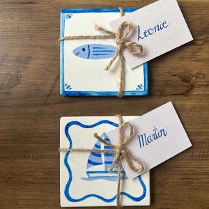 Può includere: Due sottobicchieri quadrati in ceramica con disegni nautici. Uno presenta un pesce blu, l'altro una barca a vela. Ognuno è legato con spago e ha un piccolo cartellino con un nome. Su una superficie di legno.
