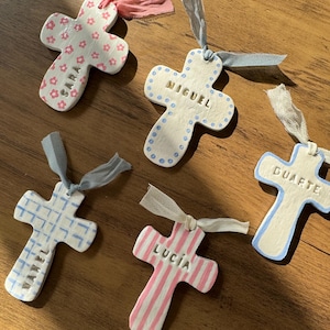 Könnte beinhalten: Handgefertigte Keramik-Kreuze mit individuellen Namen und dekorativen Mustern. Jedes Kreuz hat ein einzigartiges Design, darunter Blumen-, Streifen- und Punktmuster, und ist mit einem Band zum Aufhängen versehen. Namen wie "Sara", "Miguel", "Duarte", "Maxel" und "Lucia" sind sichtbar.