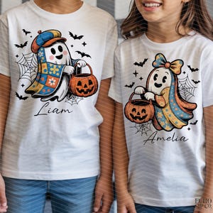 以下が含まれることがあります： ハロウィーンのゴーストデザインが施された白いTシャツ。1つのゴーストは帽子をかぶり、ジャック・オー・ランタンのバケツを持っており、下に「Liam」という名前があります。もう1つのゴーストはリボンを持ち、ジャック・オー・ランタンのバケツを持っており、下に「Amelia」という名前があります。コウモリとクモの巣も特徴です。