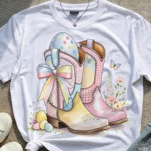 Peut inclure: T-shirt blanc avec une illustration à l'aquarelle de bottes de cowboy de Pâques. Les bottes sont rose pastel, jaune et bleu, avec des motifs floraux et vichy. Un grand nœud et des œufs de Pâques complètent le design.