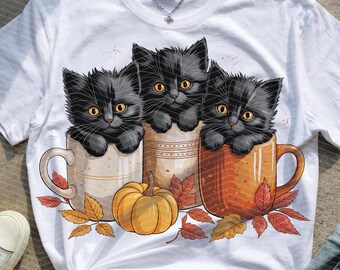 May include: White t-shirt featuring three black kittens with yellow eyes peeking out of mugs. A small pumpkin and fall leaves are between the mugs. The mugs are beige, tan, and orange.