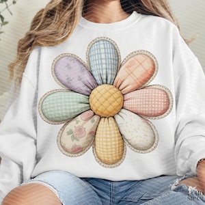 May include: White sweatshirt featuring a large, colorful flower design. The flower has quilted petals in pastel patterns and a yellow center. The design is reminiscent of a patchwork quilt, with a scalloped edge.