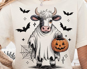 May include: Cream-colored t-shirt featuring a Halloween-themed graphic. The design includes a cow dressed as a ghost, holding a jack-o'-lantern. Black bats, stars, and a spiderweb complete the spooky illustration.