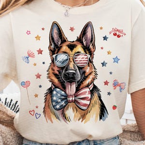 May include: Cream-colored t-shirt featuring a German Shepherd wearing sunglasses with an American flag design and a patriotic bow tie. The design includes red, white, and blue stars, bows, and hearts, celebrating Independence Day.