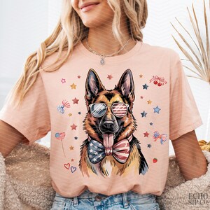 May include: A peach-coloured t-shirt featuring a German Shepherd wearing sunglasses with an American flag design and a matching bow tie. The shirt is decorated with patriotic stars, hearts, and bows.