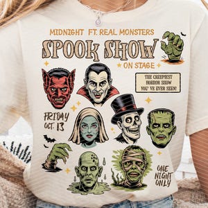 May include: Cream-coloured t-shirt with a graphic design for a "Spook Show" featuring monster illustrations. The design includes text that reads "Midnight ft. Real Monsters" and "The creepiest horror show you've ever seen!" with the date "Friday Oct. 13".