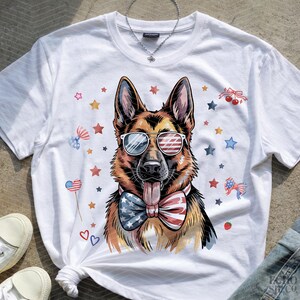 May include: White t-shirt with a German Shepherd illustration wearing sunglasses and a bow tie, both in a patriotic design. The shirt is adorned with stars and other festive decorations.