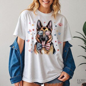 May include: White t-shirt featuring a German Shepherd wearing sunglasses with the American flag design and a bow tie with the American flag design. The dog has its tongue out. The shirt has red, white, and blue stars and other patriotic decorations.