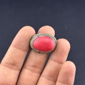 Puede incluir: Anillo ovalado con una piedra roja brillante engastada en una banda de metal plateado. El diseño del anillo es sencillo, con la piedra como centro de atención. La banda de metal tiene un aspecto texturizado, añadiendo detalles a la pieza.