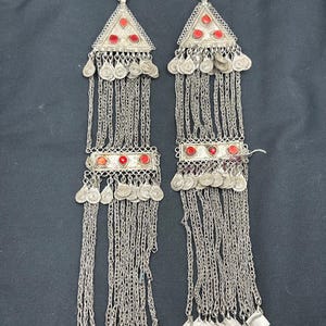 Puede incluir: Un par de pendientes de plata con piedras rojas y diseños intrincados de cadena. Los pendientes tienen una parte superior triangular con una cadena colgante de pequeños discos de plata y formas cuadradas.