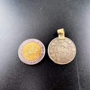 Puede incluir: Dos monedas antiguas sobre una superficie negra. Una moneda es redonda con un centro dorado y un borde morado, con una imagen y texto. La otra moneda es plateada, con un lazo dorado adjunto, y representa un relieve detallado.