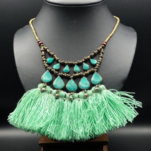 May include: A turquoise and gold-toned necklace with a mint green tassel. The necklace features teardrop-shaped turquoise stones and small beads. The tassel is made of soft, flowing threads. The necklace is displayed on a black mannequin.