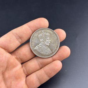 May include: A silver coin depicting a crowned profile of a man, inscribed with "EDWARD VII KING AND EMPEROR". The coin is held in a hand, with a dark backdrop highlighting the coin's features.