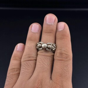 Puede incluir: Un anillo plateado con un diseño detallado que presenta un orbe central y elementos laterales ornamentados. El anillo se lleva en una mano, mostrando su intrincada artesanía y brillo metálico. El diseño del anillo incluye patrones decorativos.