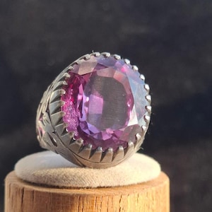 Op de afbeelding: Een zilveren ring met een grote, gefacetteerde, paarse edelsteen, gezet in een decoratieve, sierlijke zetting.