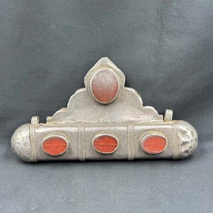 May include: A silver and red carnelian amulet with a decorative design. The amulet has a rectangular shape with a pointed top and four carnelian stones set into the front. The amulet is likely from a nomadic culture.