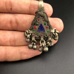 May include: A silver pendant with a triangular shape, decorated with blue, red, and green glass stones. The pendant has a fabric backing and a loop at the top for hanging. The pendant is adorned with small silver bells.