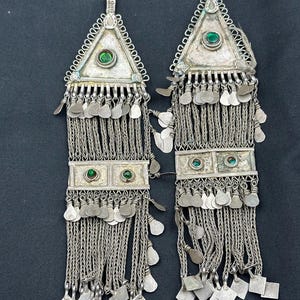 May include: Two silver earrings with a triangular top and rectangular bottom, each featuring green gemstones. The earrings have detailed chain and dangling silver accents, creating a layered, ornate design. The background is a solid black.