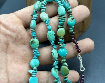 May include: A turquoise and garnet necklace with a silver clasp. The necklace is made of various sizes and shapes of turquoise stones, some of which are oval, round, and irregular. The garnet stones are small and round.