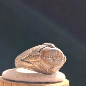 Puede incluir: Un anillo de plata con una piedra ovalada engastada en el centro. La piedra es de color marrón claro y tiene escritura árabe tallada en ella. El anillo tiene un diseño detallado alrededor de la piedra.