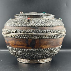 Puede incluir: Una caja redonda de madera antigua con adornos de metal plateado. La caja presenta un cuerpo de madera oscura con bandas plateadas y una tapa adornada con piedras preciosas rojas y verdes. La base y la tapa están decoradas con intrincados diseños florales y geométricos.