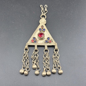 May include: A silver pendant with a triangular design, featuring red and blue gemstones. The pendant has a chain with multiple dangling balls.