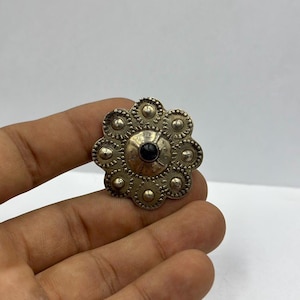 Può includere: Un fermaglio circolare in argento e nero con un centro rialzato e una pietra nera al centro. Il fermaglio è circondato da un bordo decorativo di cerchi.