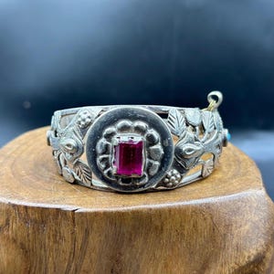 Pode incluir: Uma pulseira de prata com um design ornamentado que apresenta uma gema rosa no centro. A pulseira tem um motivo floral e é acentuada com pequenas contas.