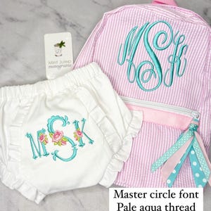 May include: A white cotton diaper with a pale aqua monogram in a master circle font. The monogram is "MSK" with pink flowers. A pink and white striped backpack with a pale aqua monogram in a master circle font. The monogram is "MSF". The backpack has a pink zipper and a pale aqua, pink, and white tassel.