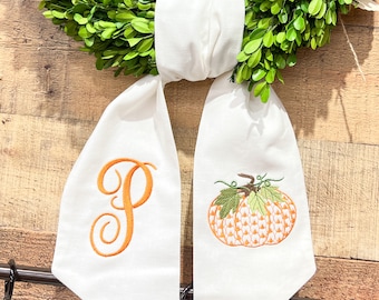 May include: White fabric ribbon with an embroidered orange pumpkin and the letter "P" in orange thread. The ribbon is tied around a green boxwood wreath.